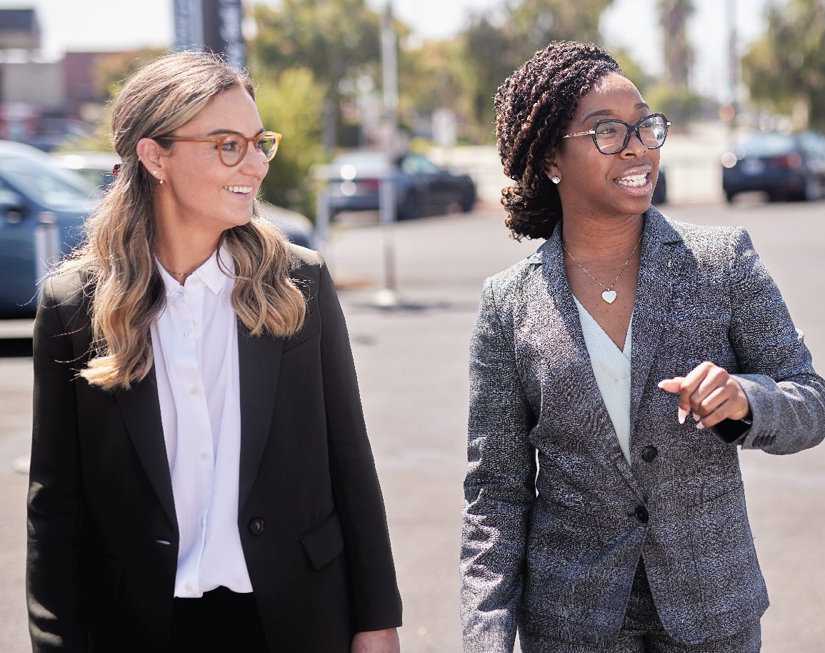 Two women in work attire talking at a car lot on a sunny day