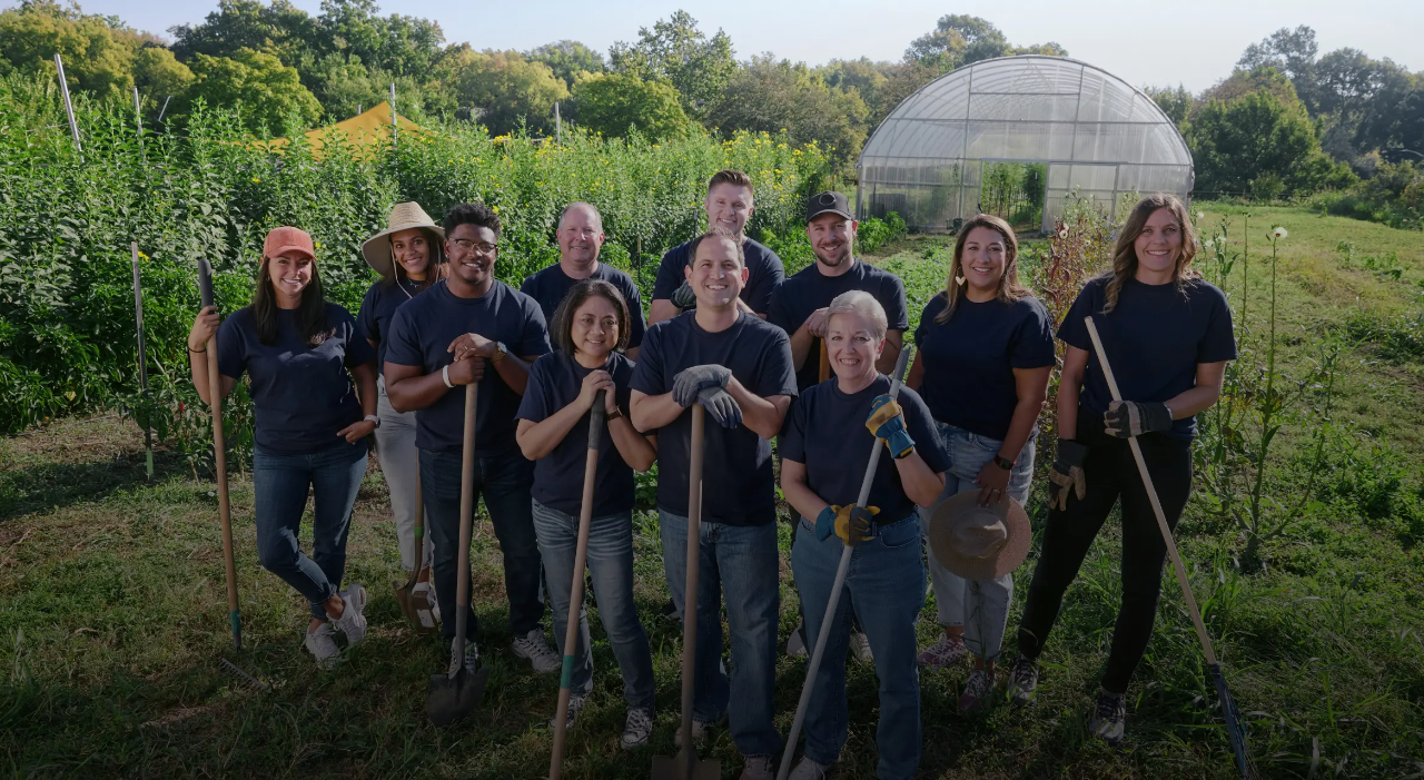 A group of smiling people holding shovels and rakes standing in a sunny field.