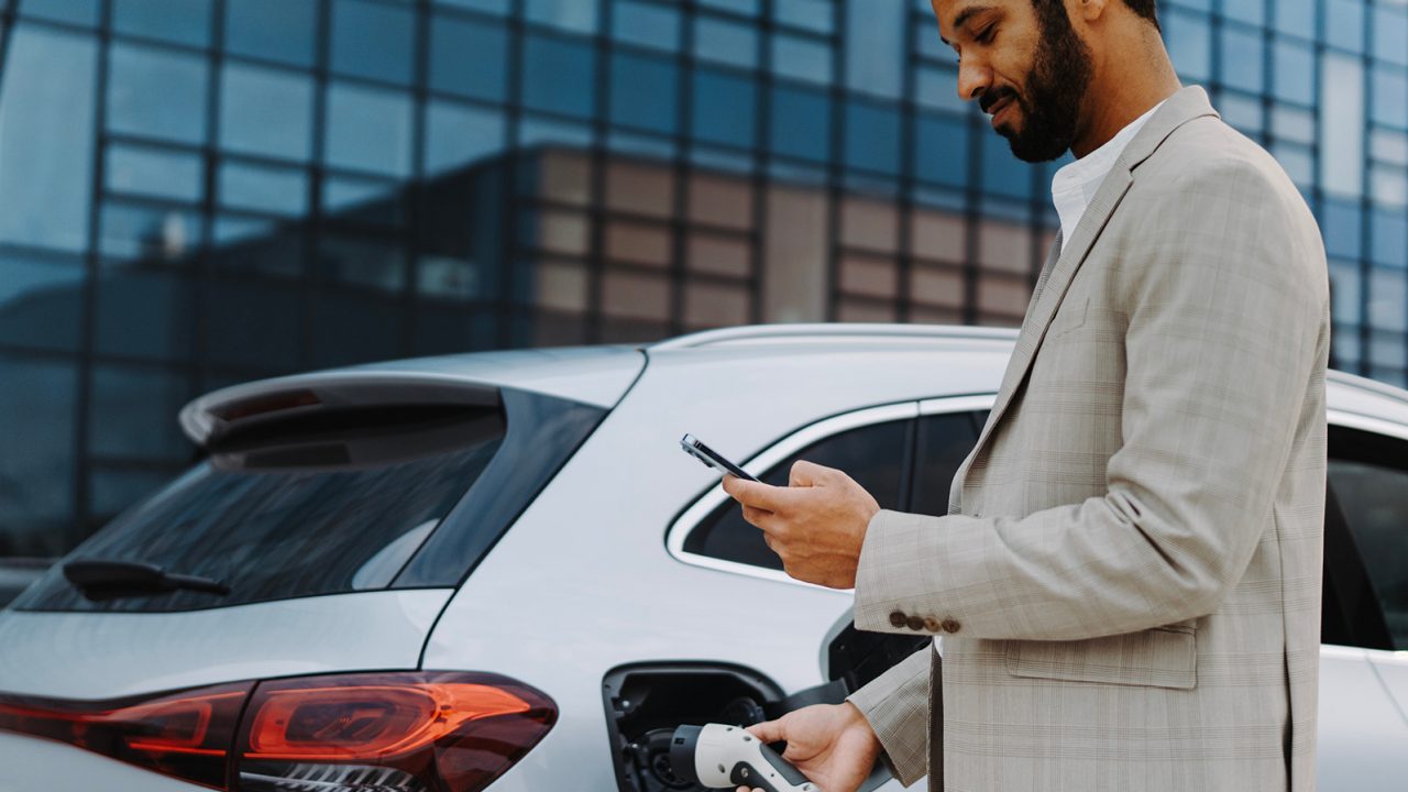 An adult male, with a beard and dress blazer, holds his cell phone and charges his electric vehicle