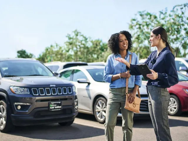 Two women talking to one another in front of a car sales lot.