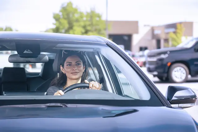 A woman behind the wheel of a blue sedan driving out of a parking lot.