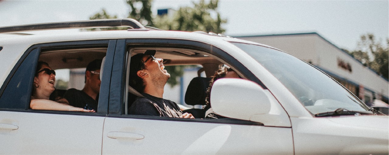 Four people laugh in a car with the windows down on a sunny day