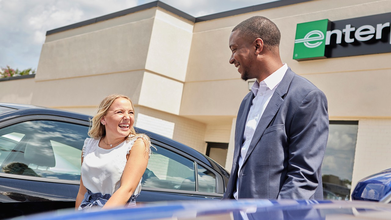 A young man and young woman, both smiling on a sunny day, talk on an Enterprise rental car lot.