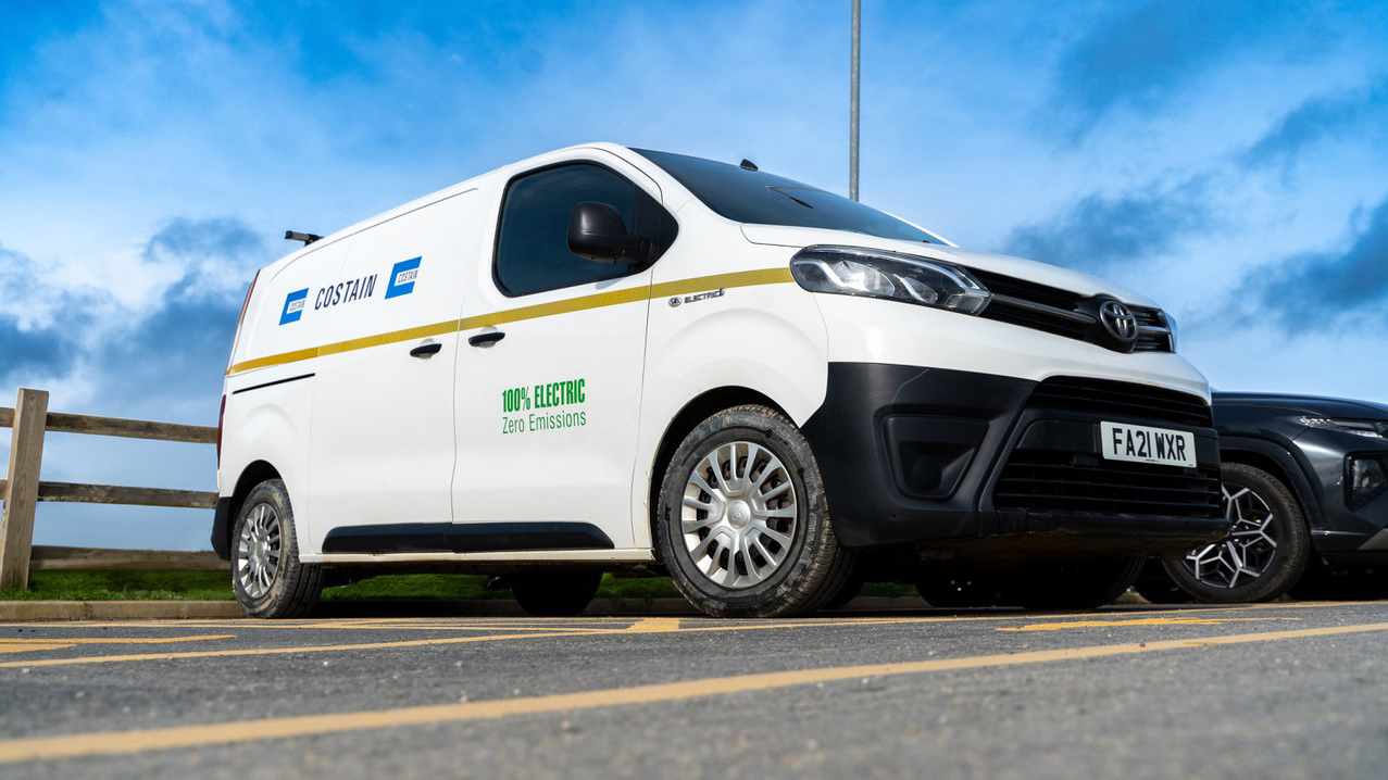 A young lady, in a seated position on green grass, leaning on a big tire and reading a book.