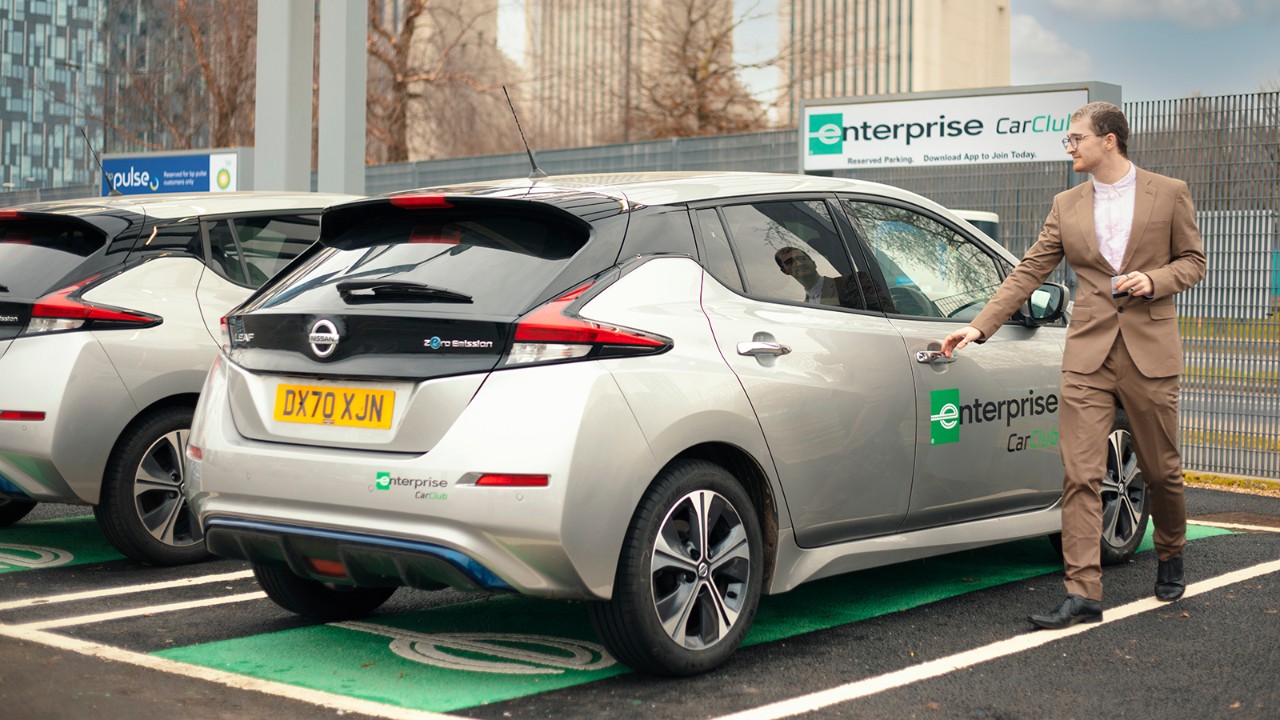 A gentleman in a tan business suit begins to open the door of a silver vehicle.