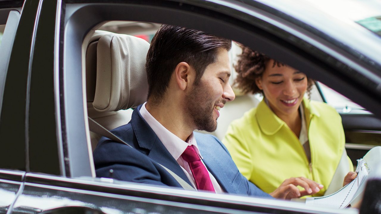 A man with a beard, shirt, neck tie and sport coat talks to a woman in a yellow shirt who is pointing at a paper. They are sitting in the front seat of a dark vehicle.