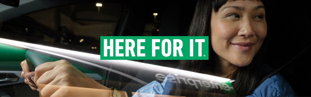 A smiling female sitting in a dark car with a "Here For It" banner as part of the graphic